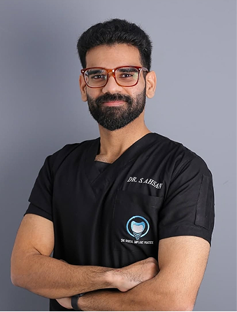 Dr. S. Ahsan, in black scrubs with dental logo, stands confidently against a gray background.