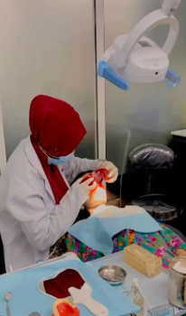 Dentist in red hijab performing a dental procedure on a patient under an overhead light in a clinic setting.